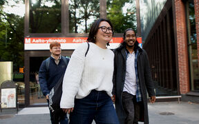 Kolme ihmistä kävelee Aalto-yliopiston metroaseman ulkopuolella. Yhdellä on valkoinen villapaita ja musta laukku.