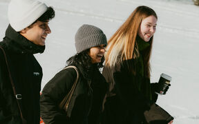Three people in winter clothing walking outdoors on a snowy day. One holds a coffee cup.