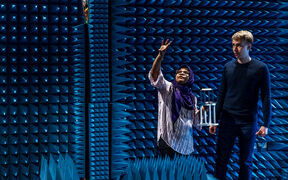 Two people in an anechoic chamber with blue acoustic foam panels. One person gestures while the other holds equipment.