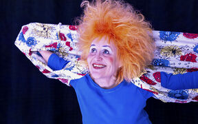 Henkilö, jolla on kirkkaan oranssit hiukset, sininen paita ja värikäs kuvioitu kangas tummalla taustalla.