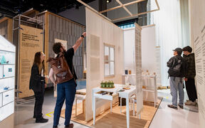 People explore a modern indoor exhibition with wooden walls and white display tables with objects.