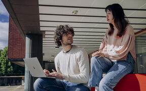 Two students outside a brick building, one using a silver laptop while talking with the other on a red bench