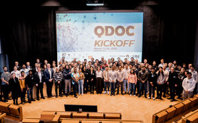 A large group of people is standing in front of a screen that reads 'QDOC KICKOFF March 11-12, 2025'.