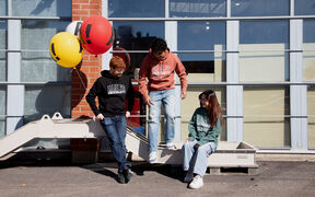 Three students in Aalto University hoodies pose outdoors by balloons and a brick wall with large windows.