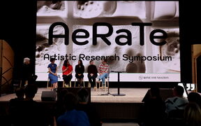 Panel of six on stage at AeRaTe Artistic Research Symposium before large abstract screen, audience watching