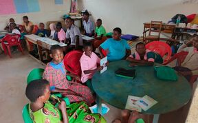 Children sit around tables in a classroom with red and green chairs, reading and talking together.