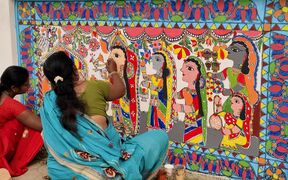 Women in bright saris painting a colourful traditional mural with stylised figures and patterns on a wall