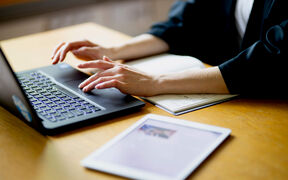 Person typing on a black laptop at a wooden desk, with a notebook and white tablet nearby
