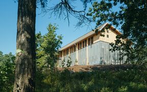 Modern light wood and metal building on a slope, surrounded by tall green trees under blue sky