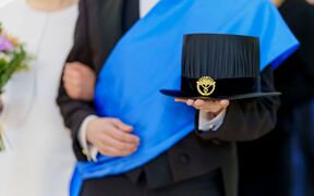 Person in formal suit with blue sash holding a black doctoral hat with a gold emblem