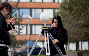 Two people outdoors adjust a tripod with a white spherical device near a brick building and trees