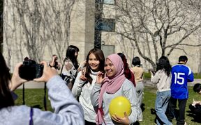 Group of students outdoors, two posing with peace signs, one holding a yellow balloon near a bare tree and building