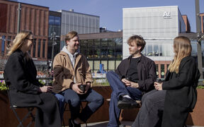 Four students sit outdoors by modern campus buildings, talking and holding coffee on a sunny day