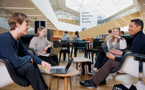 Four students are sitting on armchairs with their laptops under the sign "Better Business Better Society".