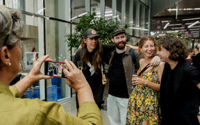 Person in green jacket photographing four friends posing indoors at a busy event
