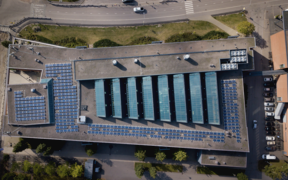Aerial view of a flat-roof building with blue solar panels and skylights, bordered by roads and trees