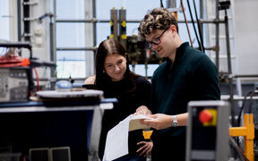 Two young adults in a lab look at papers together among machines and equipment.
