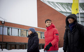 Three students in winter coats walk in snow by a red brick campus building with a yellow flag.