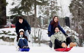 Four friends in winter coats sledding downhill on snow, laughing in front of a forest and cabins