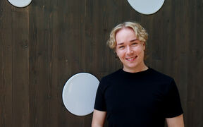 Smiling Arttu Jääskeläinen with blond curly hair in a black T-shirt, standing by a dark wooden wall with round white panels.