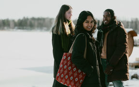 Three friends in winter coats stand smiling outdoors on snow, one with a red patterned tote bag.