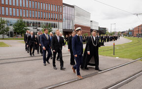 Henkilöitä tummissa puvuissa kävelemässä kampuksella raitiovaunukiskojen yli. Taustalla Väre-rakennus.