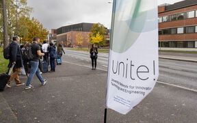 Ihmiset matkatavaroiden kanssa odottavat kampustien varrella Unite!-lipun ja syyspuiden lähellä.