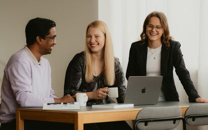 Three people having a discussion around a table with a laptop, notebook, and coffee cups.