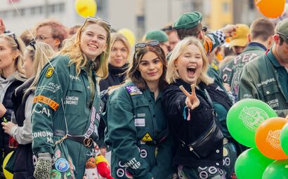 Students in green overalls at a lively outdoor event, posing for photos among orange and green balloons