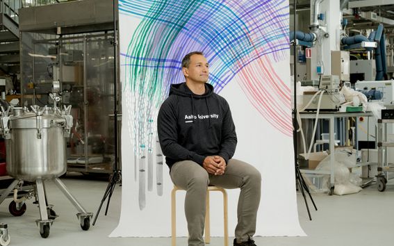 Michael Hummel istumassa laboratoriossa. Taustalla on kuvitettu kangas, jossa näkyy koivun runkoja. Kuvaaja: Rainer Paananen. Kuvittaja: Satu Kettunen.