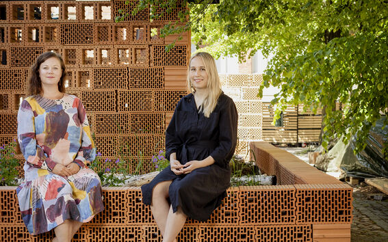 Kaksi naista istuu tiilistä tehdyn muurin päällä. Toisella on päällä musta mekko, toisella kukikas.