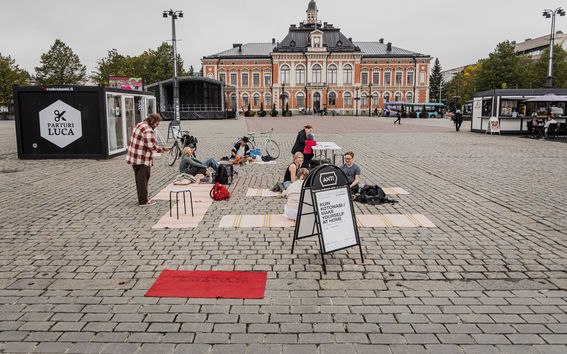 Six people sitting on the ground on the carpets and at a table in the middle of the city. In the background is a large orange building, Kuopio City Hall.