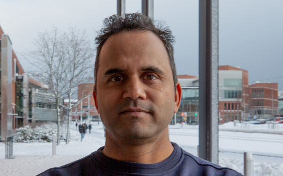 Ashish Thapliyal in a dark jumper stands indoors with a snowy urban campus scene through the window behind.
