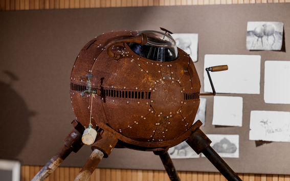 A rusty, spherical metal contraption with tripod legs and a glass window, displayed indoors with sketches on the wall.