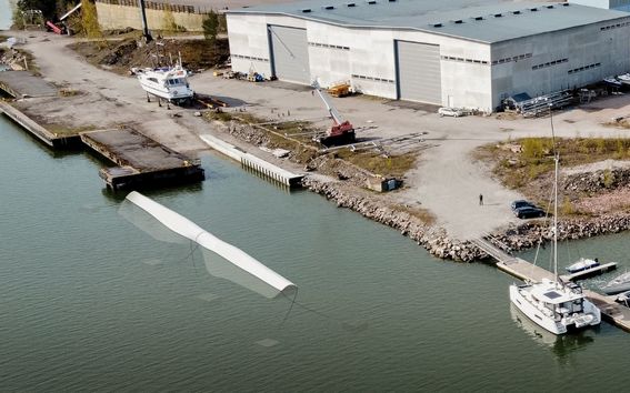 Ilmakuva suuresta rakennuksesta veden äärellä, jossa on veneitä laiturissa ja tuulivoimalan siipi vedessä aallonmurtajana.