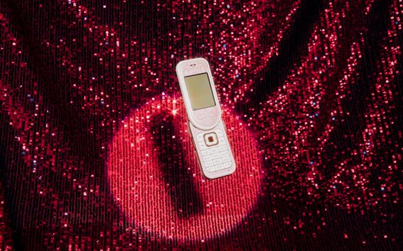 A white flip phone with a screen open lies on a sparkling red sequined fabric.