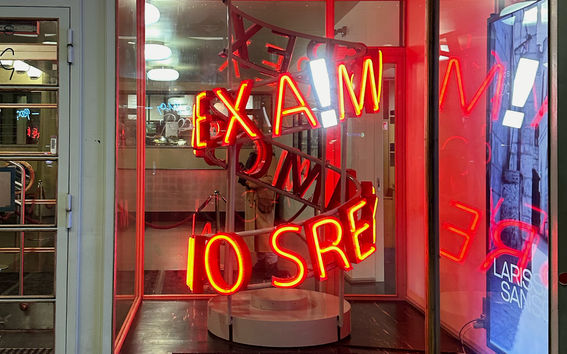 Ett skyltfönster med en röd neonskylt som lyder 'EXA!M 10 SRE'. Reflektioner syns på glaset.