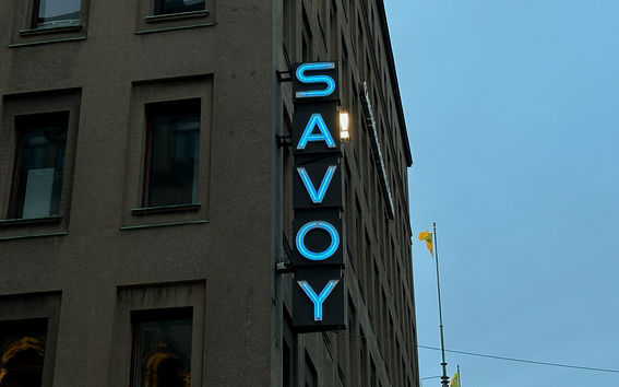 Skylt för 'Savoy' med blåa neonljus på hörnet av en grå byggnad med fönster. Gult flagg på stång i närheten.