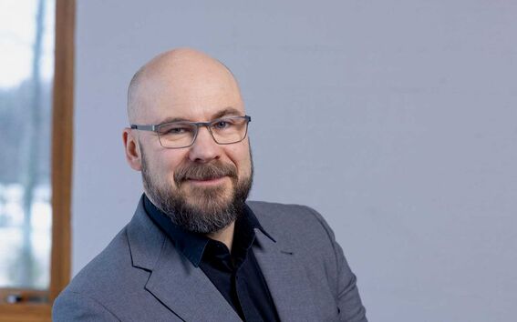 Jaan Praks, jolla on harmaa puvun takki ja musta paita, istuu puisella ikkunanpuitteilla varustetun ikkunan edessä.