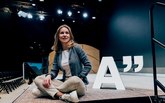 Person sitting cross-legged on a stage next to a large 'A' and quotation mark sign, under stage lighting.
