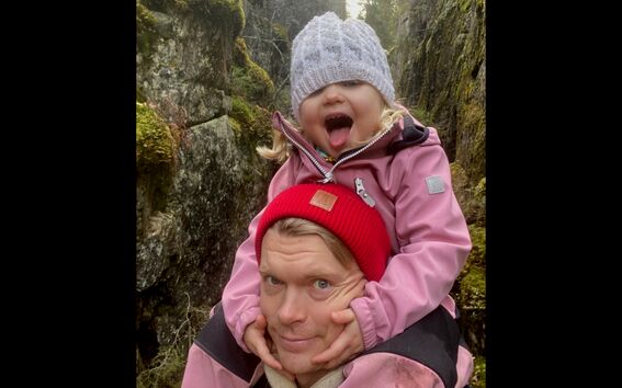 A child in a pink jacket and grey hat hugs another in a red beanie. They are outdoors with moss-covered rocks behind them.