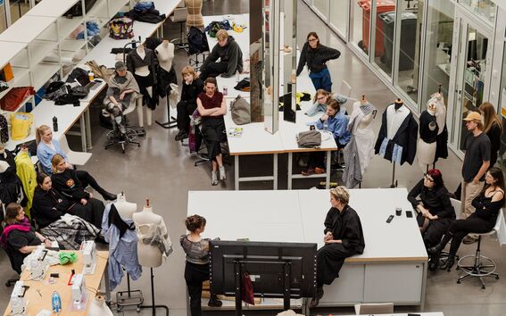 A group of people are gathered in a fashion design studio with mannequins, sewing machines, and fabric materials.