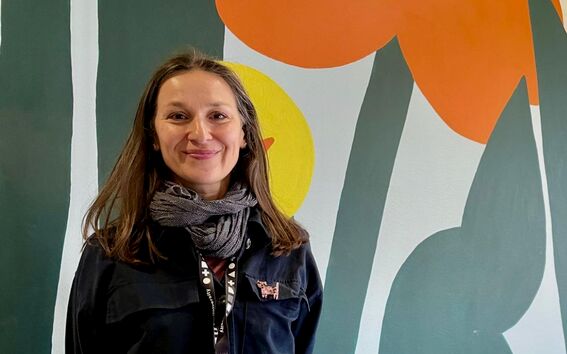 Person wearing a black jacket and grey scarf, with a conference badge. Background has flower murals in green, orange and yellow.