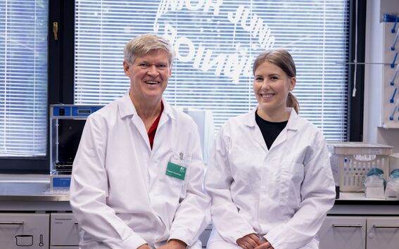 Two people in white lab coats sitting at a table in a laboratory with a window in the background.