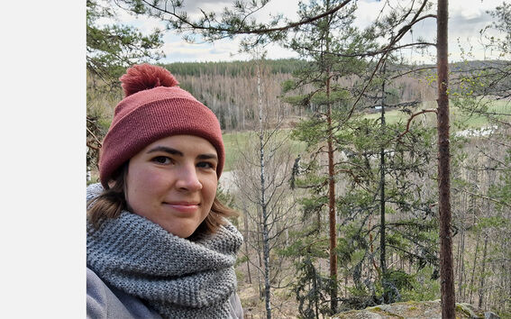 Punaisen hatun ja harmaan huivin pukeutunut henkilö seisoo mäellä ja katsoo metsään laaksoon mäntyjen ympäröimänä.