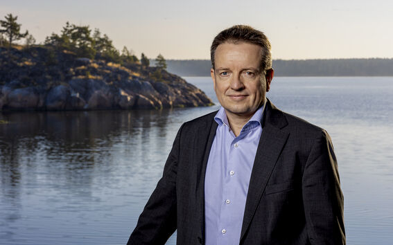 A man in a grey suit stands by a calm lake with forested islands in the background.
