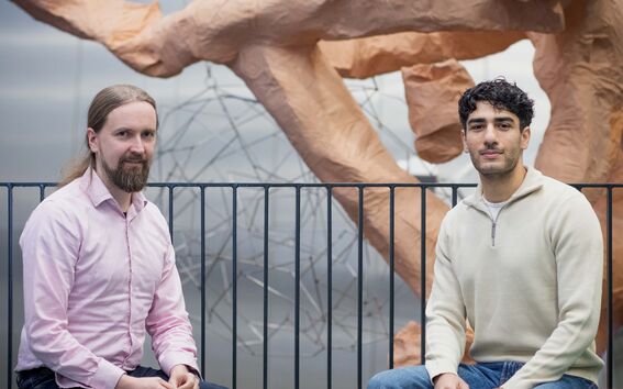 Two men sitting in front of a large abstract sculpture made of brown material, with a metal fence behind them.