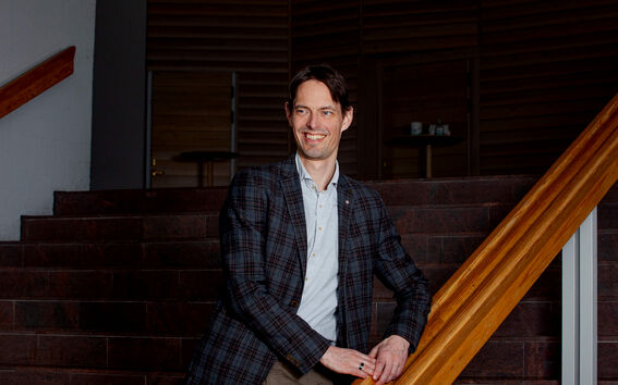 A person in a plaid jacket and light shirt stands by a wooden railing on a staircase indoors.