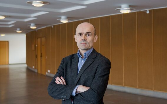 A person in a grey blazer stands with arms crossed in a hallway with wooden panelled walls and ceiling lights.