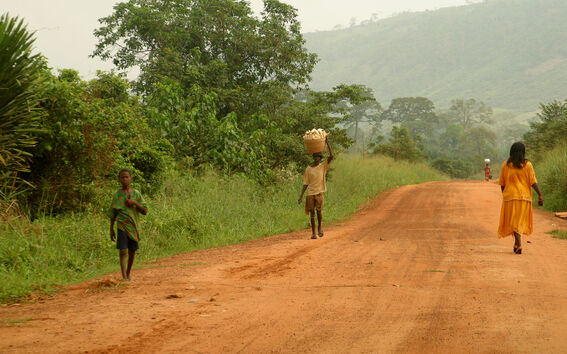 Soratie, jolla kävelee ihmisiä. Yksi henkilö kantaa koria päänsä päällä. Taustalla vihreitä puita ja kukkuloita.